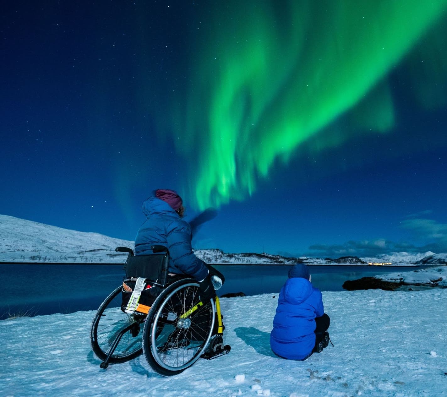 Una persona en silla de ruedas y su acompañante, en la nieve, miran una aurora boreal verde en el cielo de la noche.