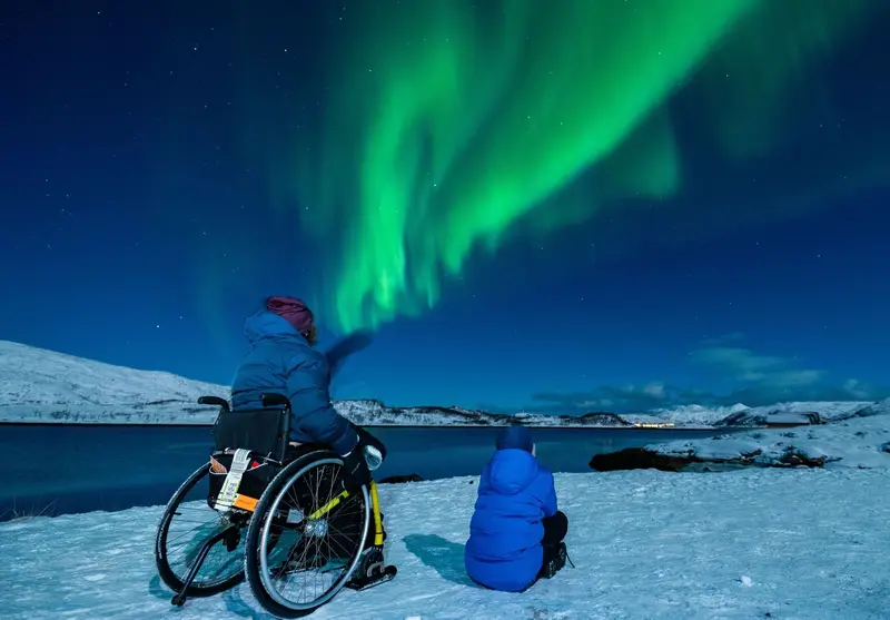 Usuaria de silla de ruedas y su hijo sentado en la nieve divisando auroras boreales en Tromso, noruega