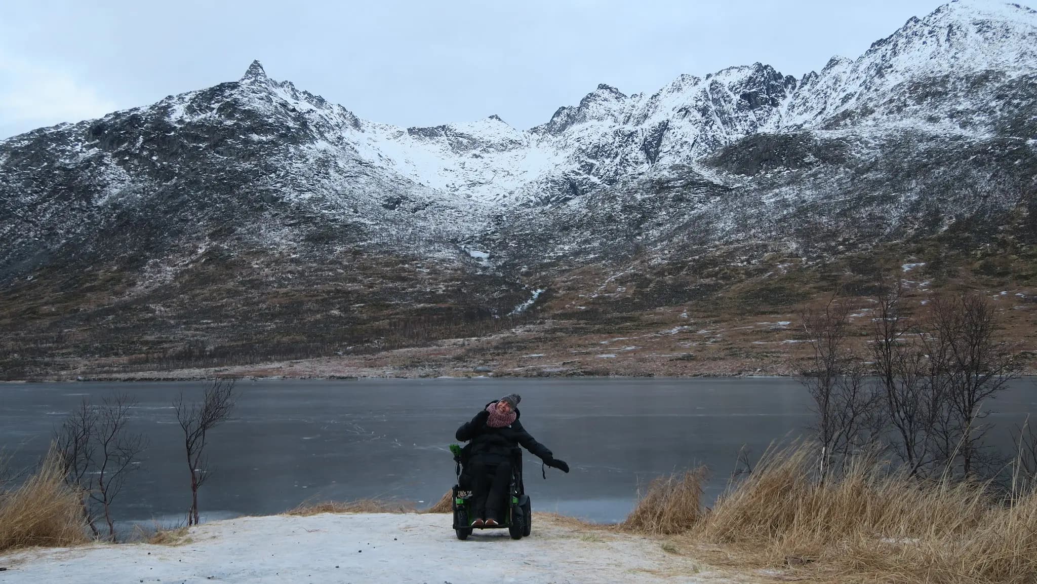 Una persona en silla de ruedas eléctrica saluda a la orilla de un lago helado, entre montañas nevadas, en Noruega.