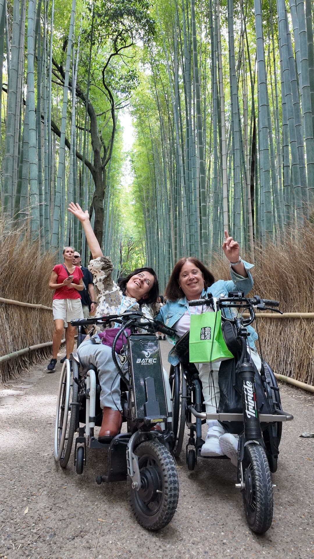 Dos viajeras en silla de ruedas con handbike levantan los brazos, sonrientes, en un bosque de bambú.