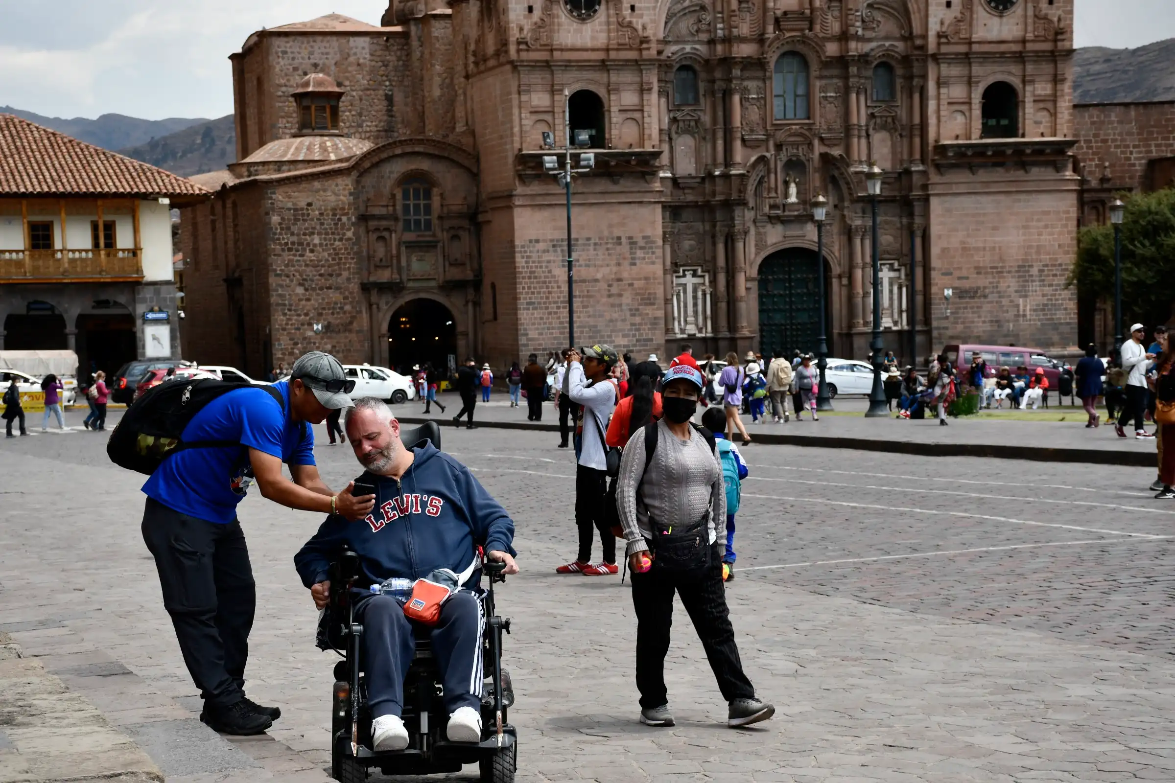 Un viajero en silla de ruedas eléctrica, con un acompañante que se agacha a su lado, en la plaza de Armas de Cusco, en Perú.