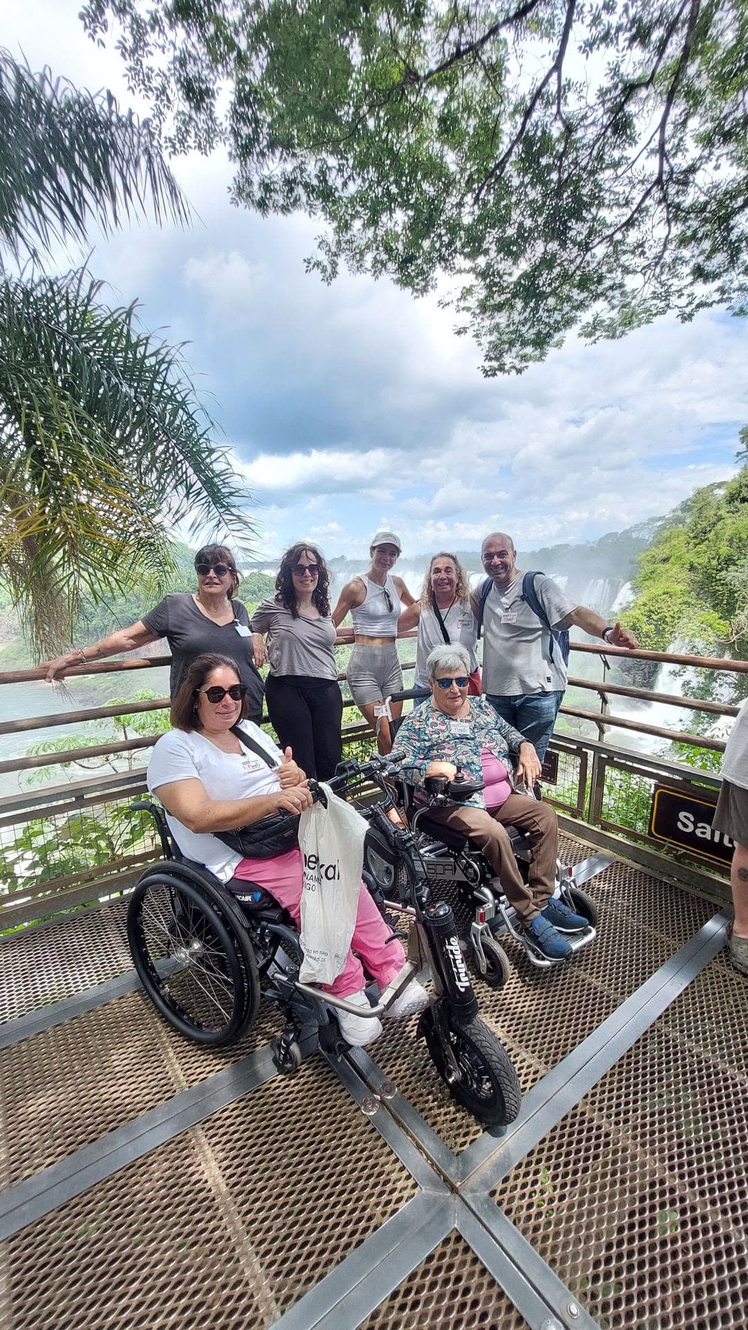 Un grupo de viajeros, dos en silla de ruedas, sonríe en una pasarela metálica rodeada de selva.