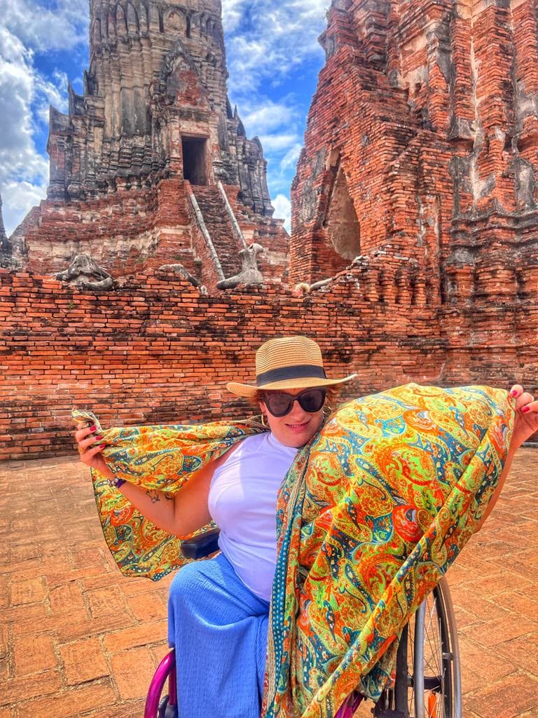 Una viajera en silla de ruedas extiende un pañuelo de colores delante de un templo de ladrillo rojo.