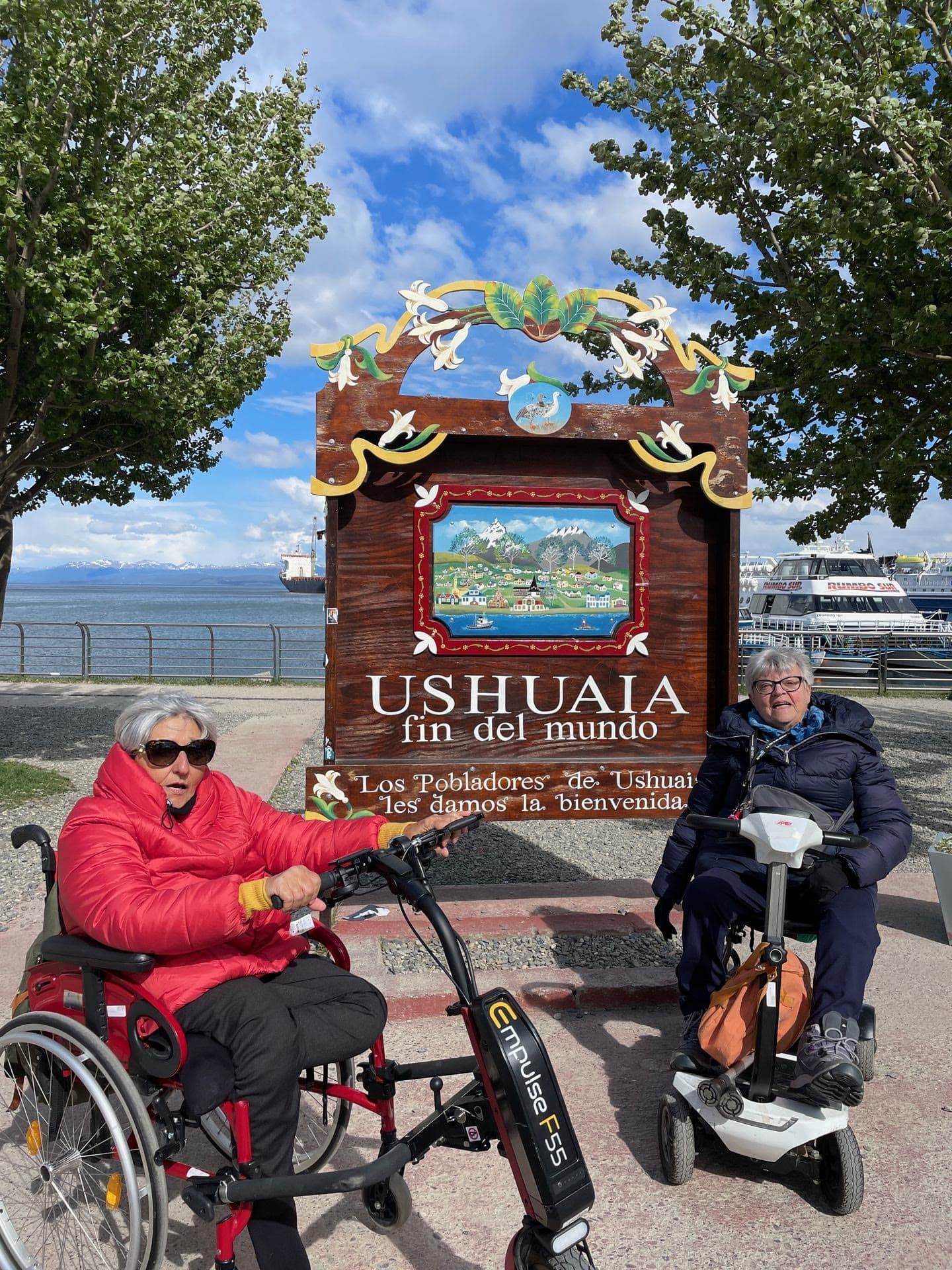 Dos viajeros, uno en silla de ruedas y otro en scooter, junto al cartel «Ushuaia, fin del mundo».