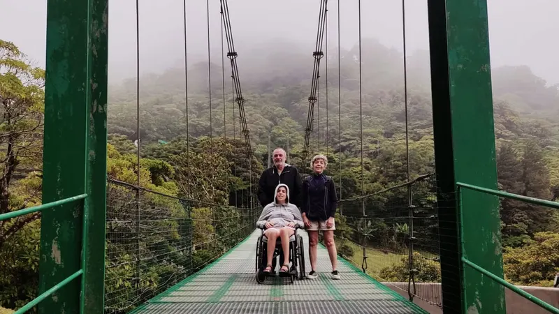 Tres miembros de una familia, la hija usuaria de silla de ruedas, cruzando un puente colgante en Costa Rica. Hay un ambiente nuboso.