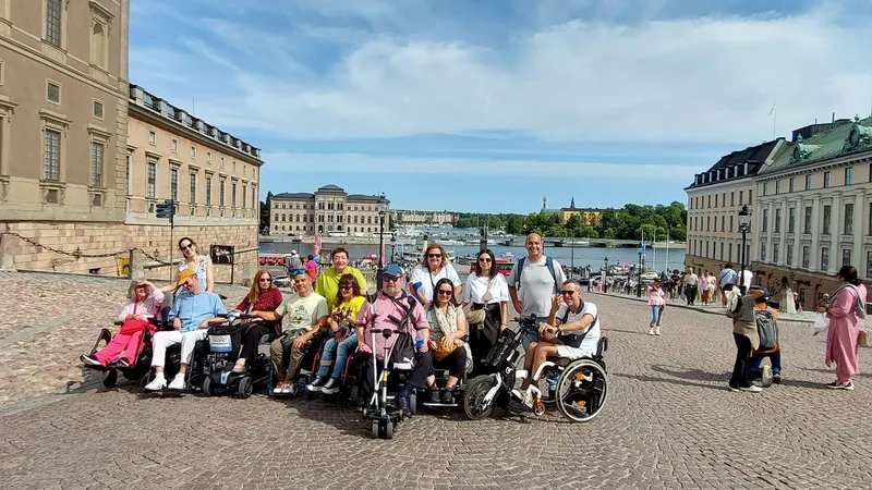 Un grupo de personas usuarias de silla de rueda junto con 2 acompañantes en una calle de Estocolmo. Al fondo se puede ver el mar y un edificio histórico de la ciudad.
