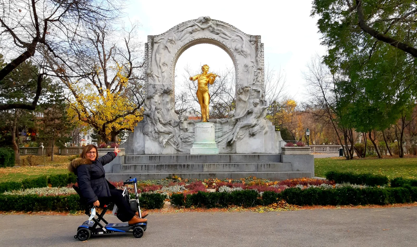Persona en scooter en parque frente a monumento clásico con arco de piedra y estatua dorada, paseo pavimentado.