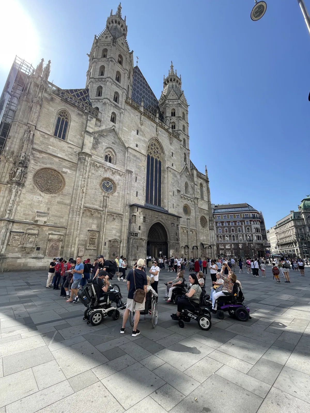 Catedral con fachada gótica de piedra blanca y plaza de suelo plano y pavimentado donde se ven personas en sillas de ruedas y cochecitos de bebé.