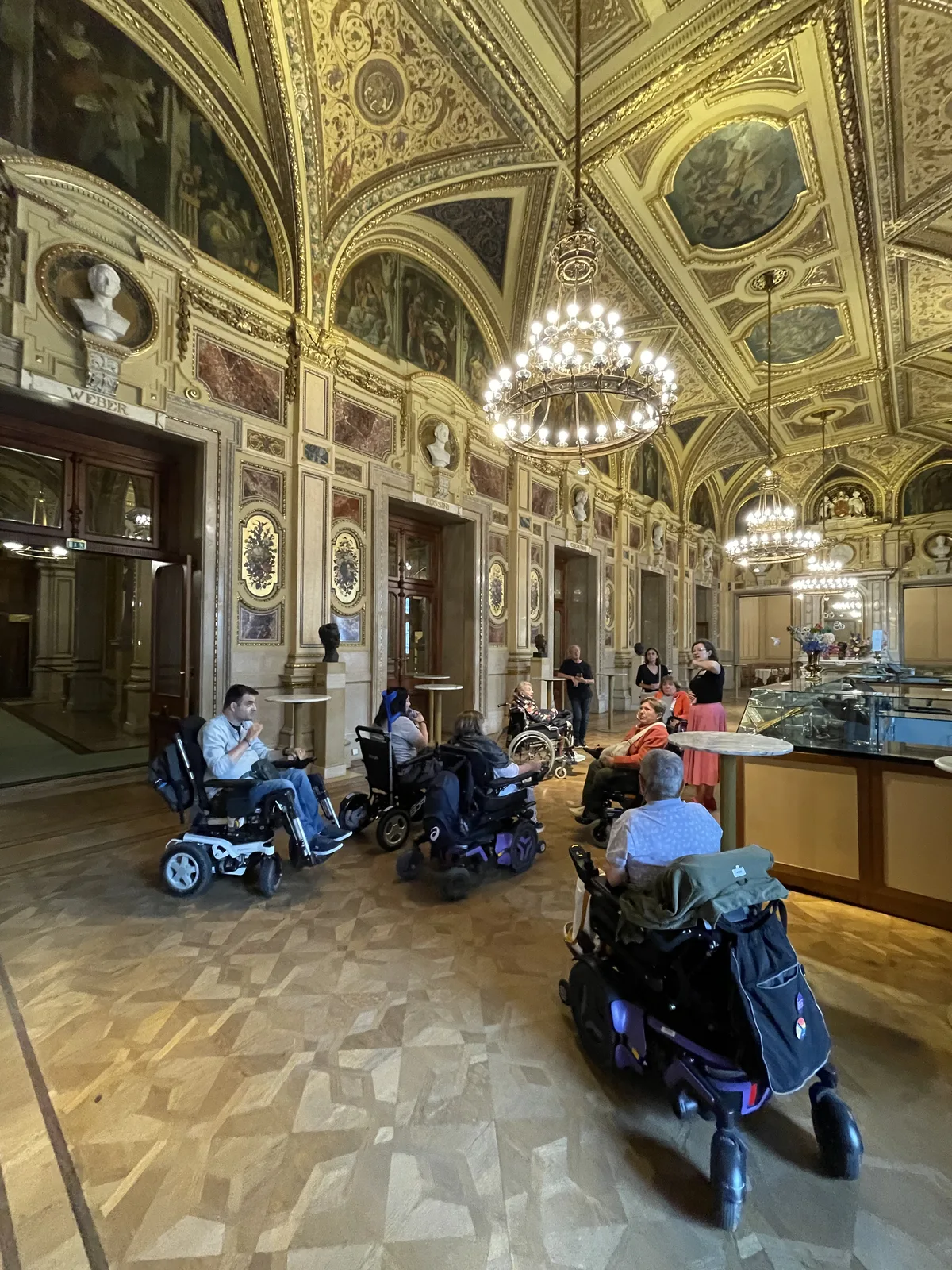 Interior de palacio con ornamentación dorada, frescos en techos abovedados, lámparas de araña, suelo de mosaico y personas en sillas de ruedas motorizadas azules en el espacio.