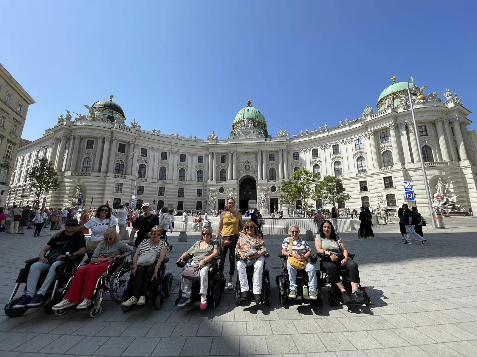 Plaza con edificio histórico de arquitectura clásica. Varios visitantes con sillas de ruedas y personas de pie en primer plano. Pavimento plano y despejado.