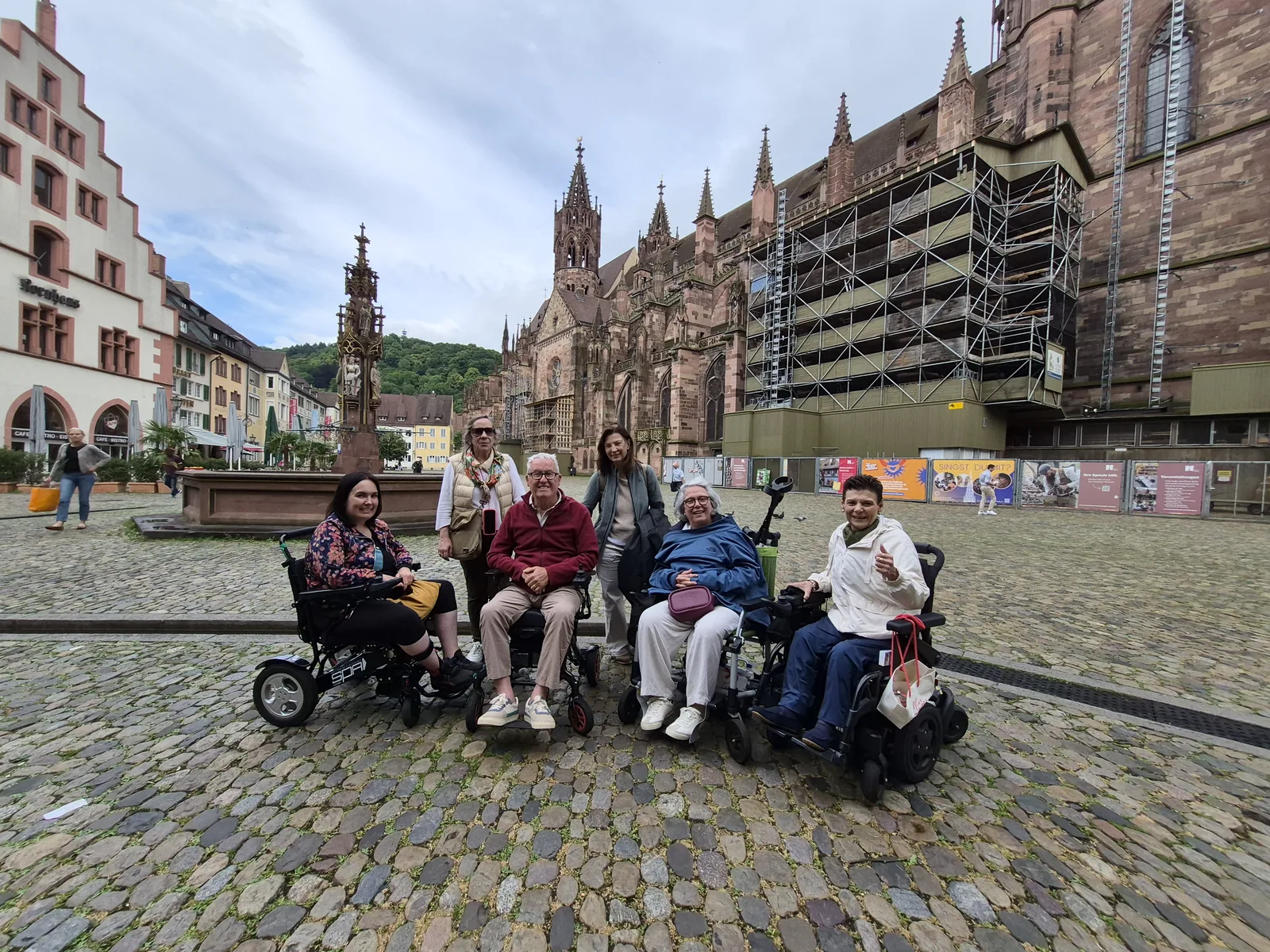 Plaza empedrada con catedral gótica al fondo. Grupo de personas en sillas de ruedas y de pie posando para foto. Edificios históricos y andamios de restauración visibles.