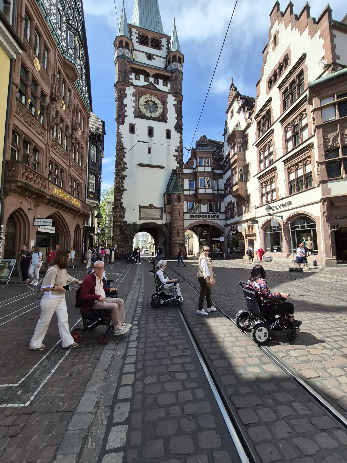 Plaza medieval con torres históricas, pavimento plano de adoquines, vías de tranvía y personas caminando.