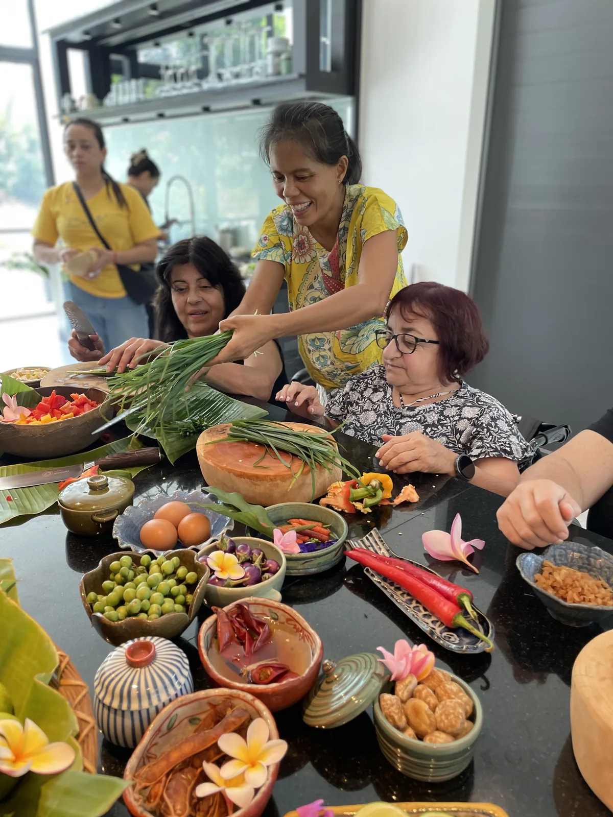 Grupo de personas en taller culinario alrededor de mesa con frutas y verduras frescas, ambiente interior moderno con mostrador de cocina al fondo.