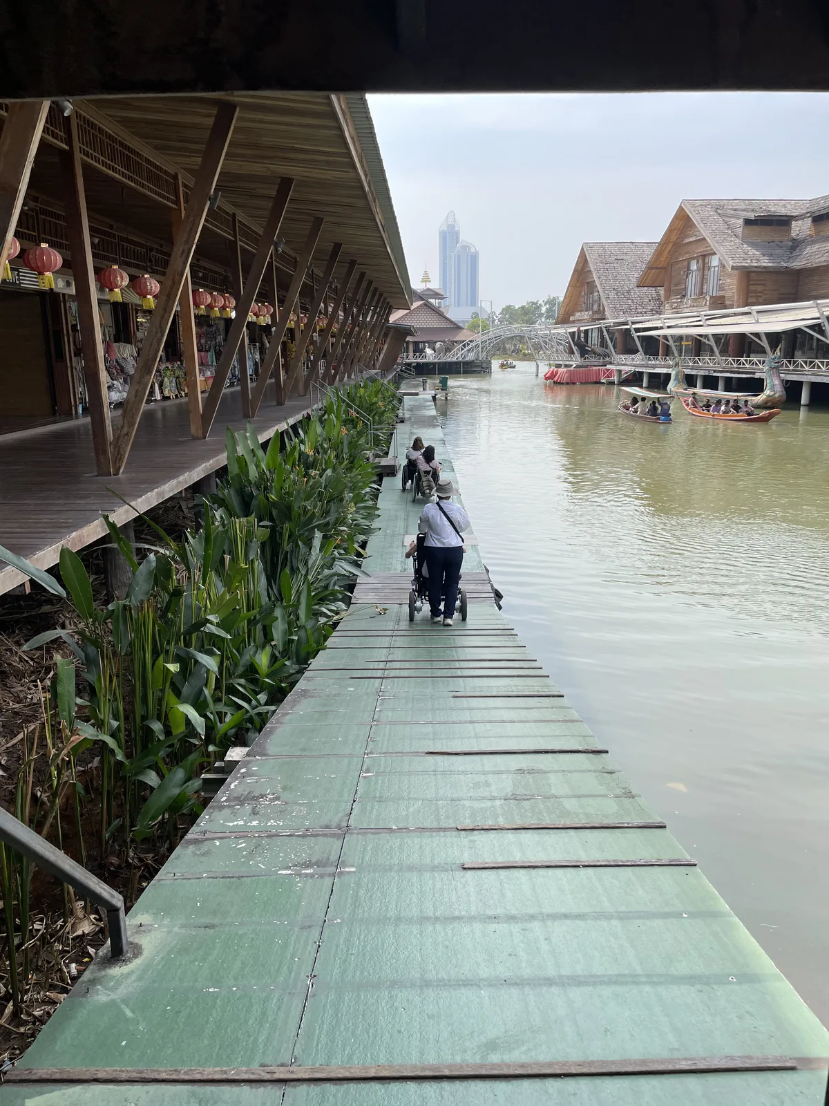 Pasarela de madera junto a edificios tradicionales en canal de agua. Persona en silla de ruedas circulando por la plataforma elevada. Entorno turístico con casas flotantes y vegetación acuática.