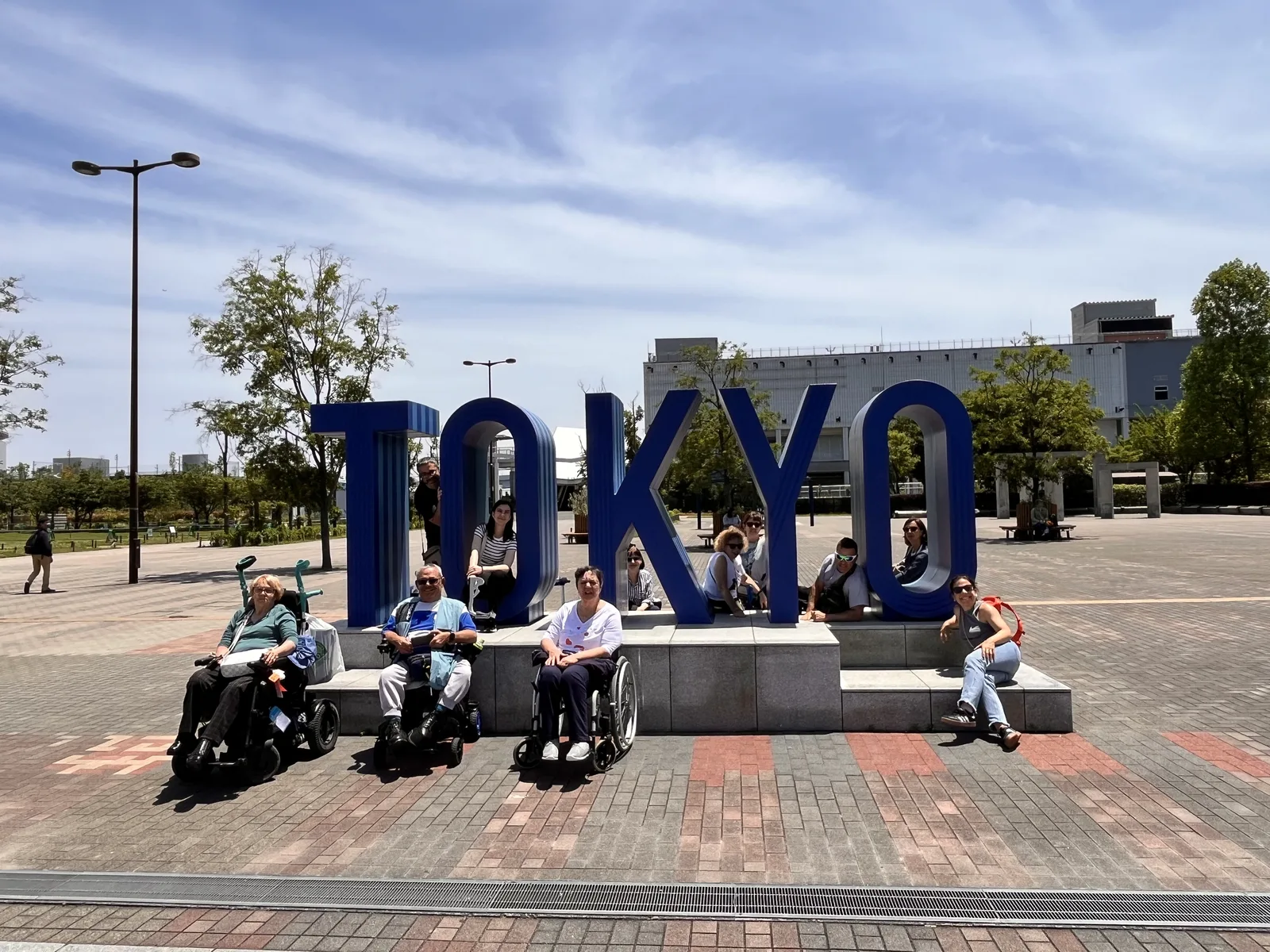 Plaza con piso pavimentado plano, monumento de letras grandes, personas en sillas de ruedas frente al letrero TOKYO, árboles y edificios de fondo.