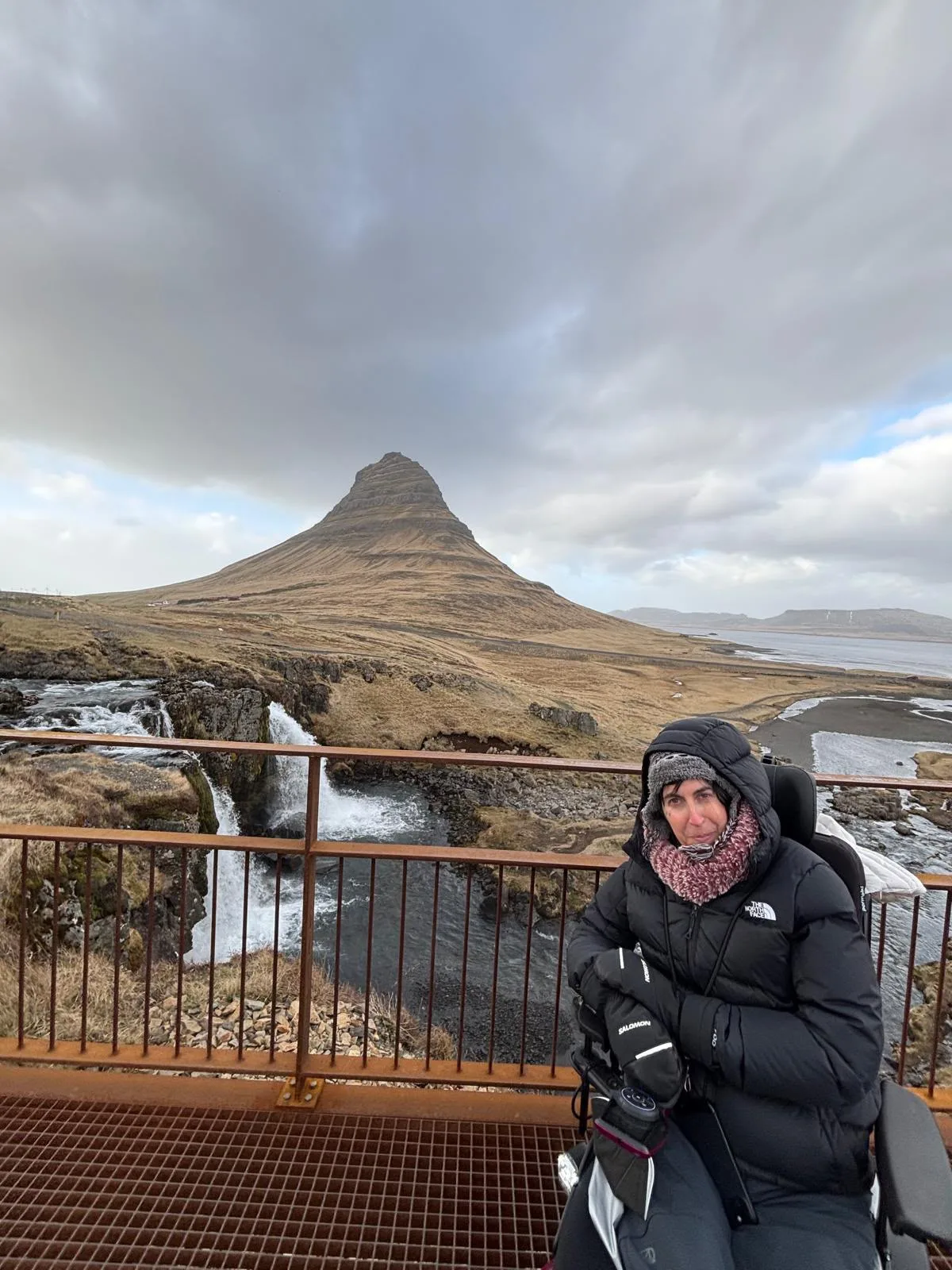 Persona con abrigo negro en una plataforma de madera elevada con baranda metálica, observando una montaña cónica bajo cielo nublado. Pasarela con vista panorámica de paisaje islandés con cascada.