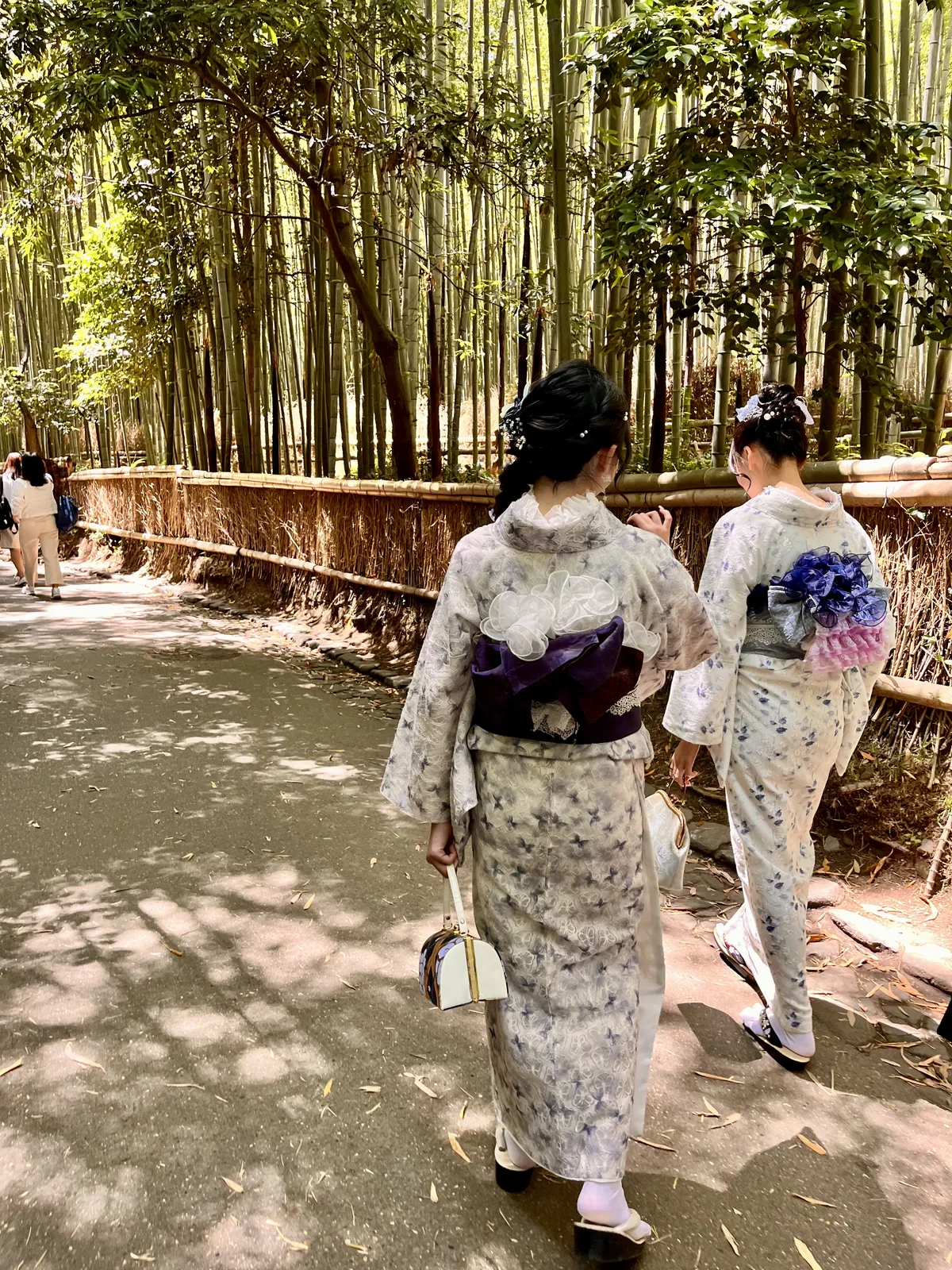 Pasarela de madera en bosque de bambú. Dos personas caminan de espaldas vistiendo kimonos tradicionales. Vegetación densa a ambos lados, luz natural filtrada.