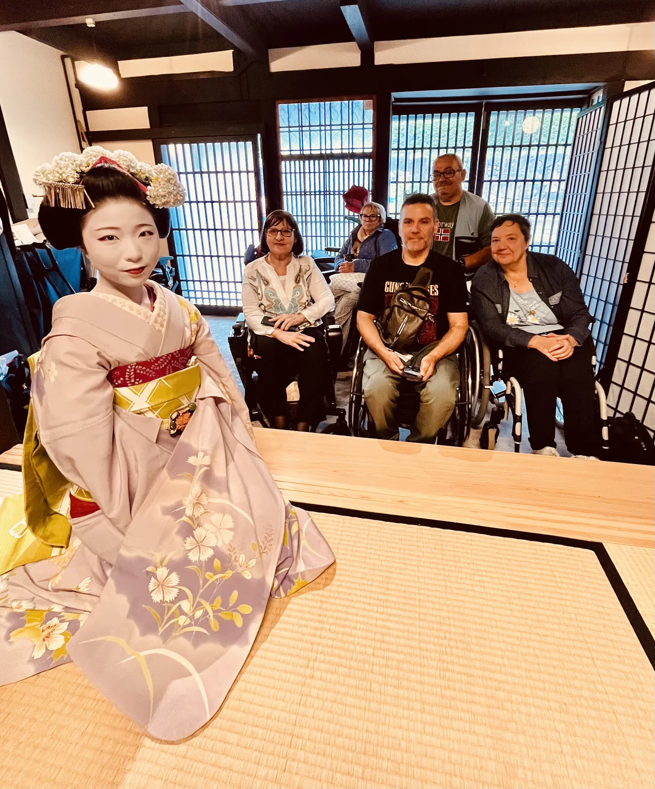 Interior de una casa tradicional japonesa con tatami. Una persona con kimono en primer plano. Al fondo, un grupo de personas, una de ellas en silla de ruedas, sentadas en el suelo observando. Puertas correderas de papel tradicionales al fondo.