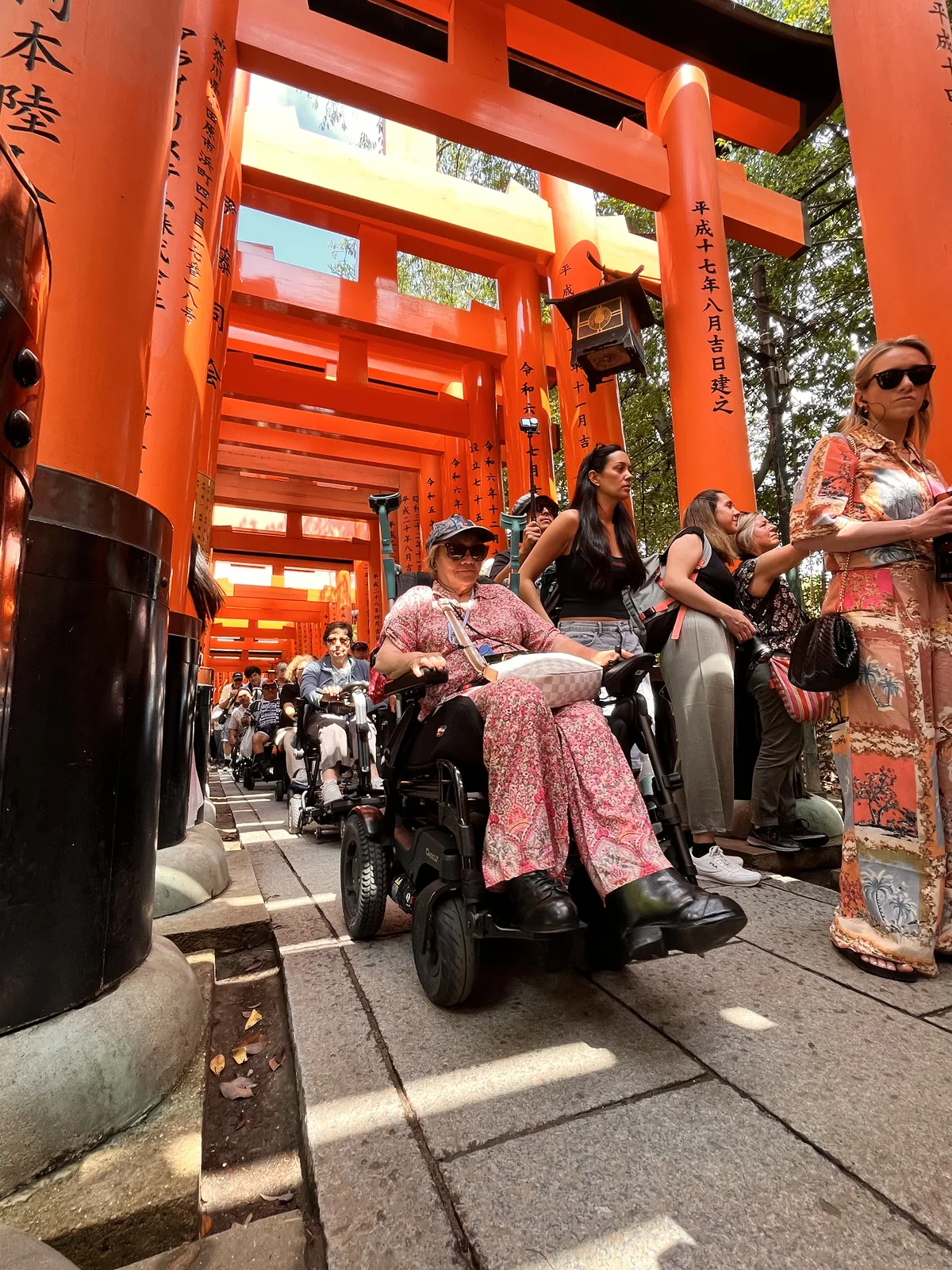 Pasaje con torii rojos tradicionales japoneses. Una persona en silla de ruedas manual avanza por el camino pavimentado entre los pórticos. Ambiente de templo o santuario con visitantes alrededor.