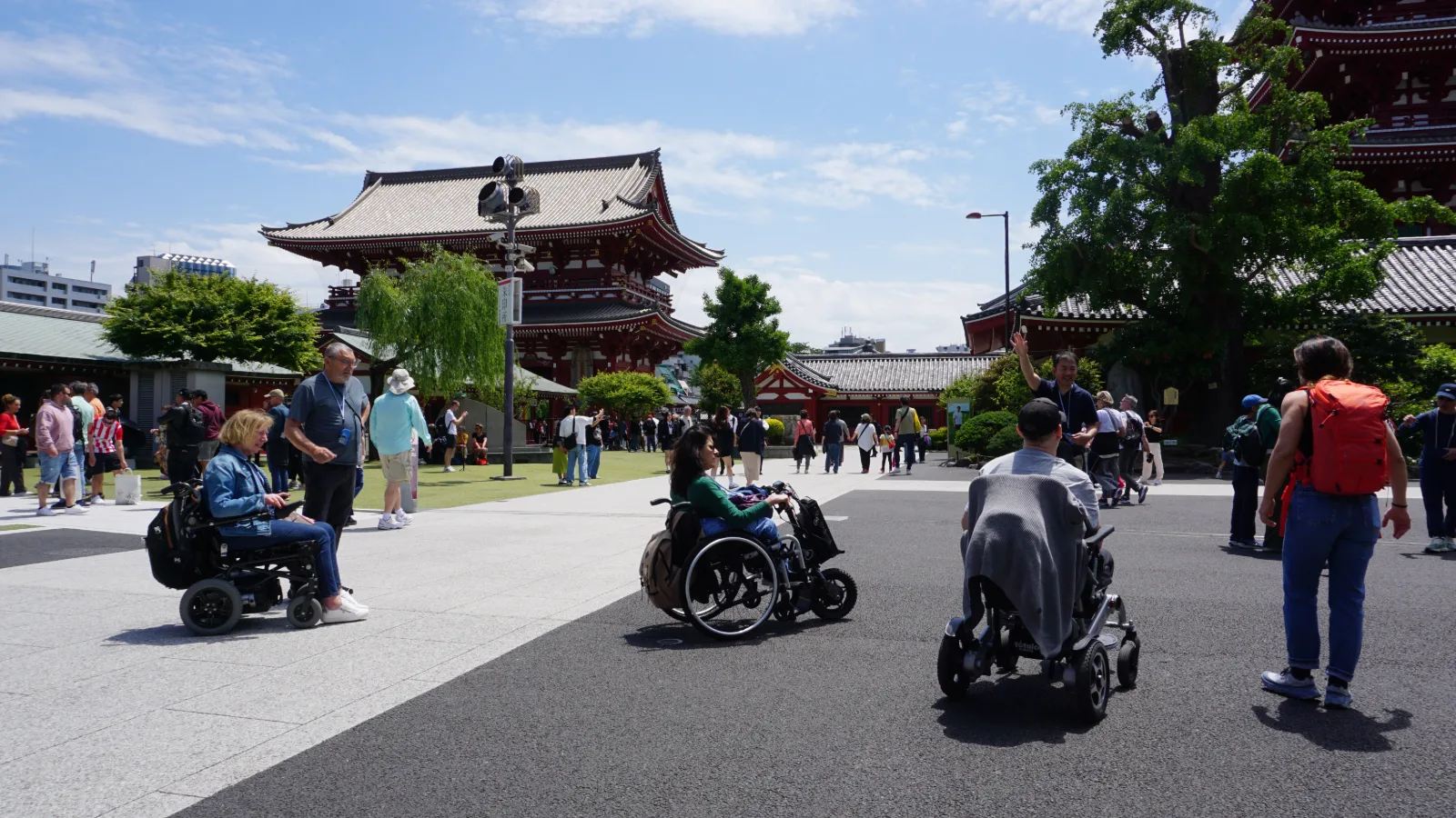 Templo budista tradicional japonés con tejado curvo rojo oscuro y detalles decorativos. En la plaza accesible frente al templo, varias personas en sillas de ruedas y con movilidad reducida disfrutan del espacio. Árboles verdes rodean el complejo, y multitud de visitantes caminan entre los edificios