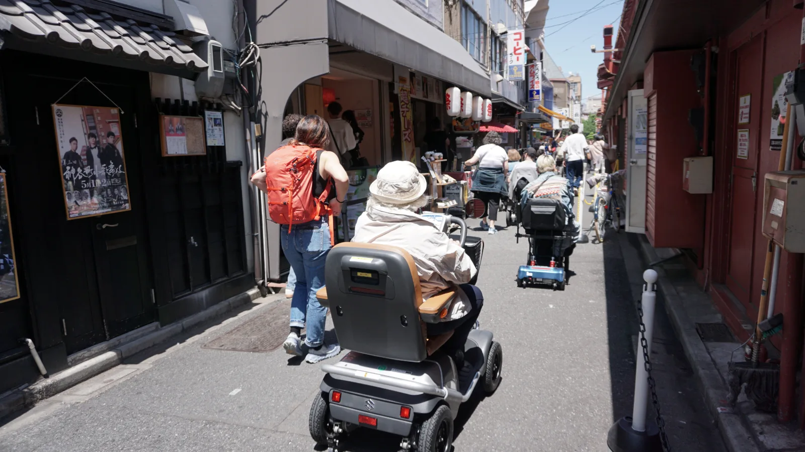 Calle estrecha de un barrio tradicional en Japón con tiendas a ambos lados. Varias personas con cochecitos de bebé y carros de compras pasean bajo el sol. Una mujer en primer plano lleva una mochila roja. Faroles de papel rojo cuelgan sobre los negocios. El pavimento es accesible y la calle está bie