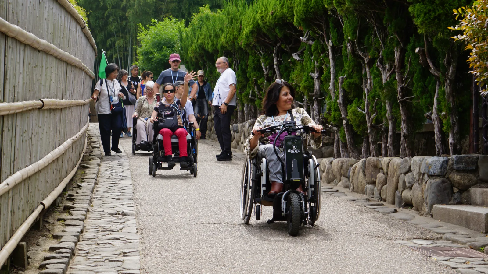 En un camino empedrado entre muros de piedra y cipreses, una mujer en silla de ruedas motorizada roja y otra persona en triciclo adaptado avanzan junto a un grupo de visitantes durante un recorrido accesible por un sitio histórico en Japón.