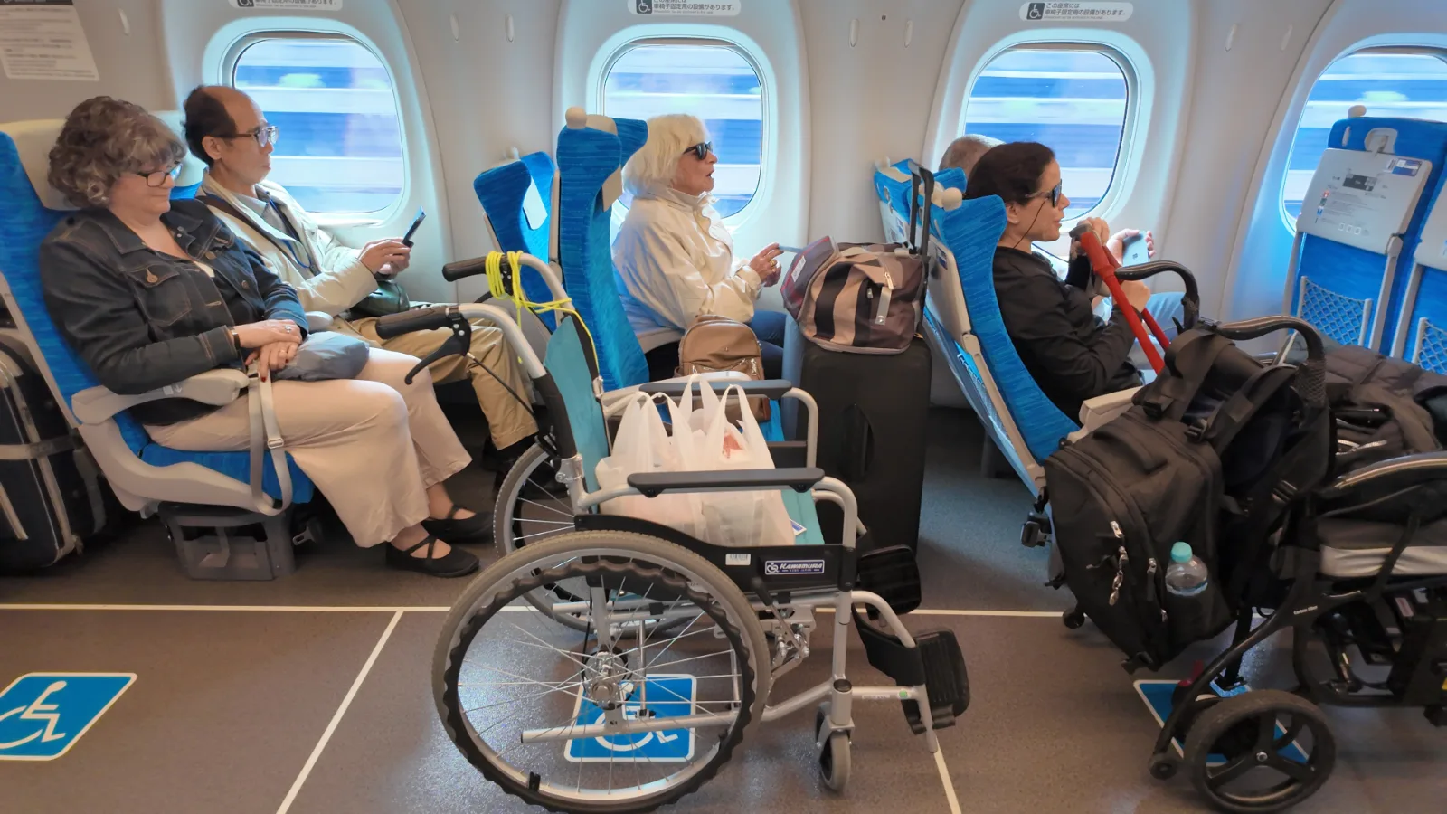 Interior de avión con varios pasajeros en sus asientos. En primer plano, sillas de ruedas estacionadas en el pasillo. Ventanillas redondas muestran el cielo azul. Asientos azules y grises. Señalización de accesibilidad visible en el suelo.