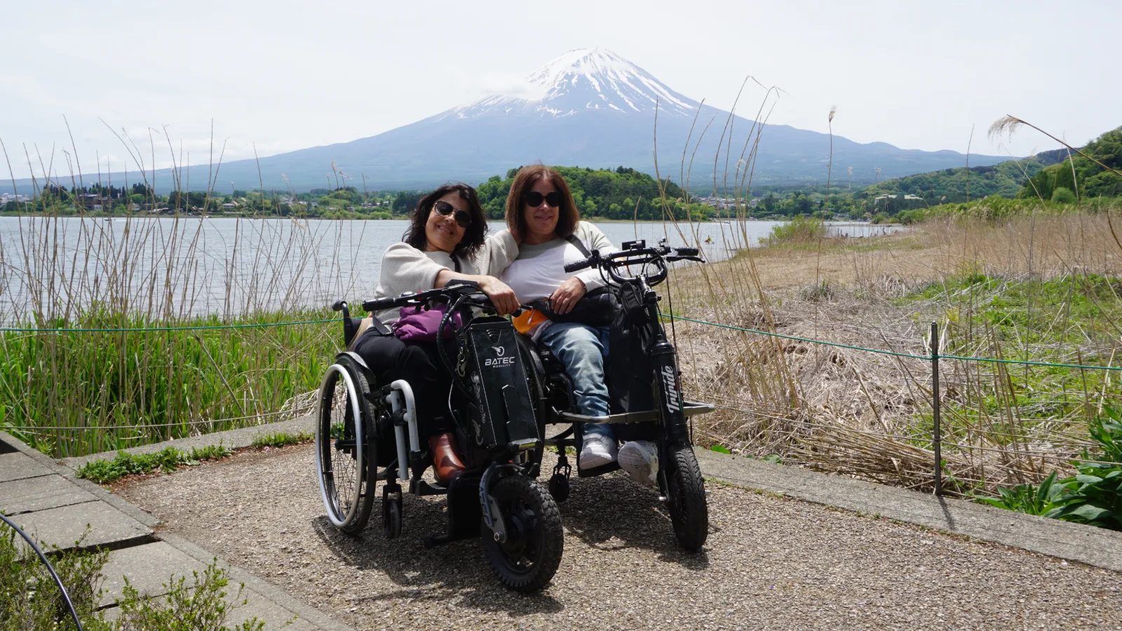 Dos mujeres en un triciclo adaptado con silla de ruedas posan frente a un lago. Al fondo, el monte Fuji nevado domina el paisaje. La orilla está rodeada de vegetación acuática y plantas verdes.