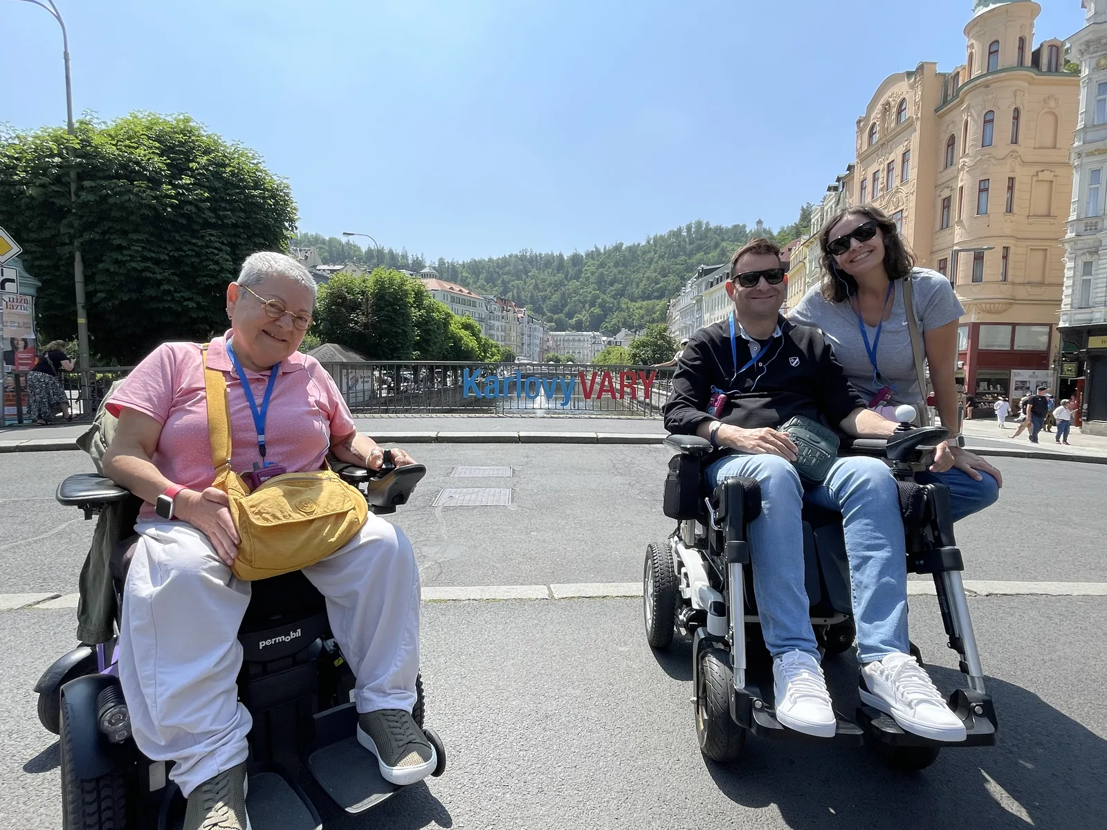 Dos mujeres en sillas de ruedas motorizadas en una plaza pública con edificios históricos y montañas al fondo.