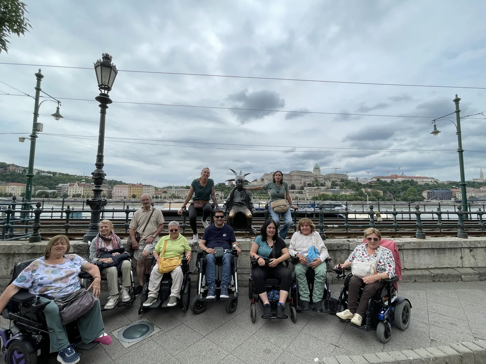 Grupo de personas en sillas de ruedas y de pie en una plaza peatonal junto a un río, con farolas ornamentales y casas en las colinas al fondo.
