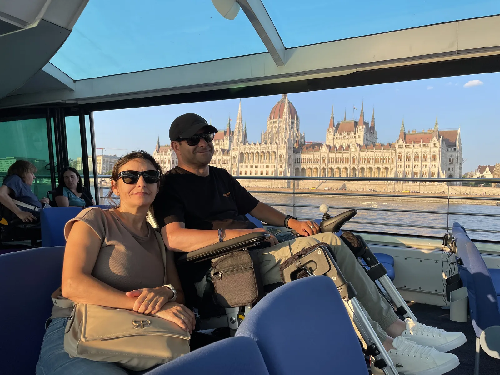 Pareja en barco turístico con vista al Parlamento de Budapest y el río Danubio. Cubierta con asientos, barandillas y amplio espacio de circulación.