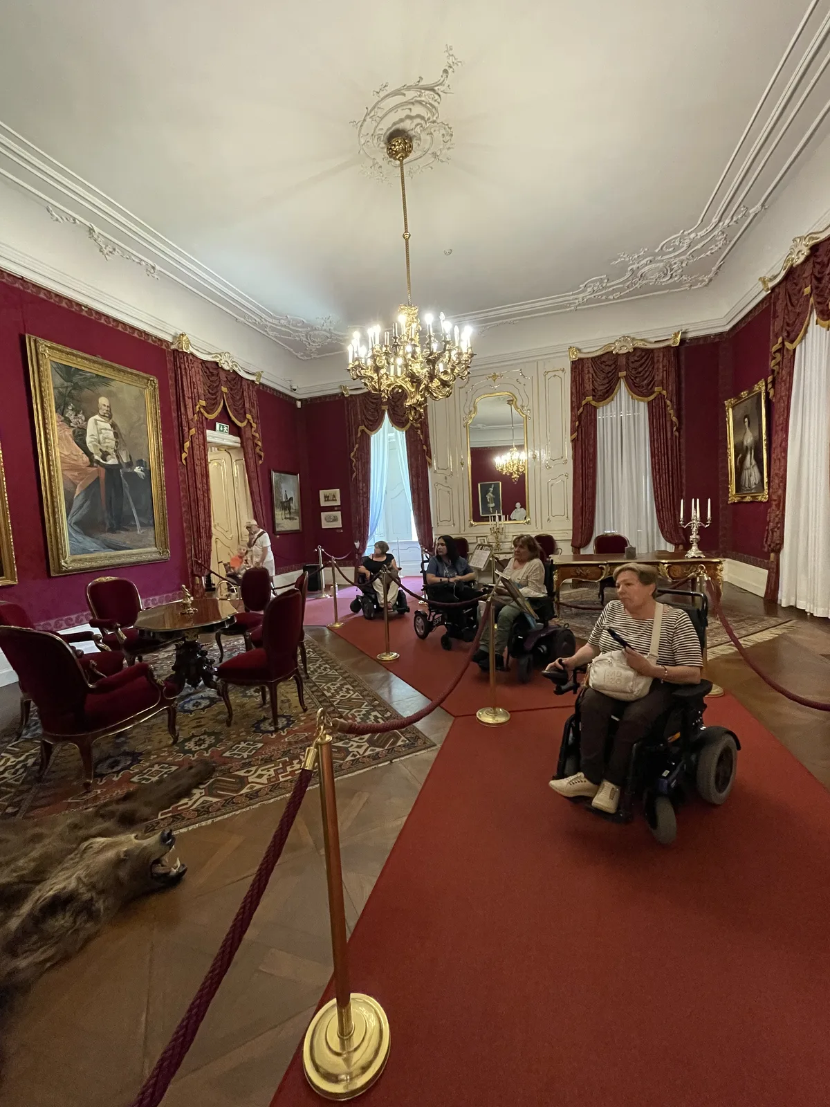 Sala elegante de palacio con paredes granate, lámpara de araña, cuadros al óleo y una persona en silla de ruedas circulando por pasillo con suelo rojo.